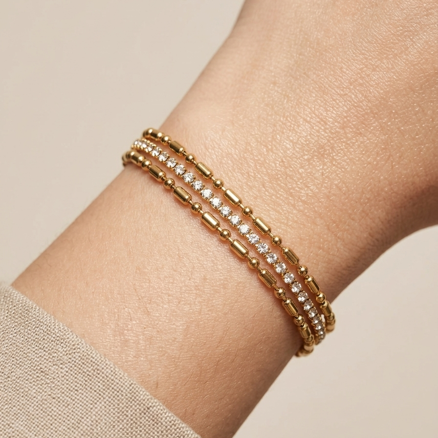 Close up of a wrist wearing a layered 18k gold plated bracelet featuring delicate gold beads and a row of sparkling CZ diamonds wrist against a beige background
