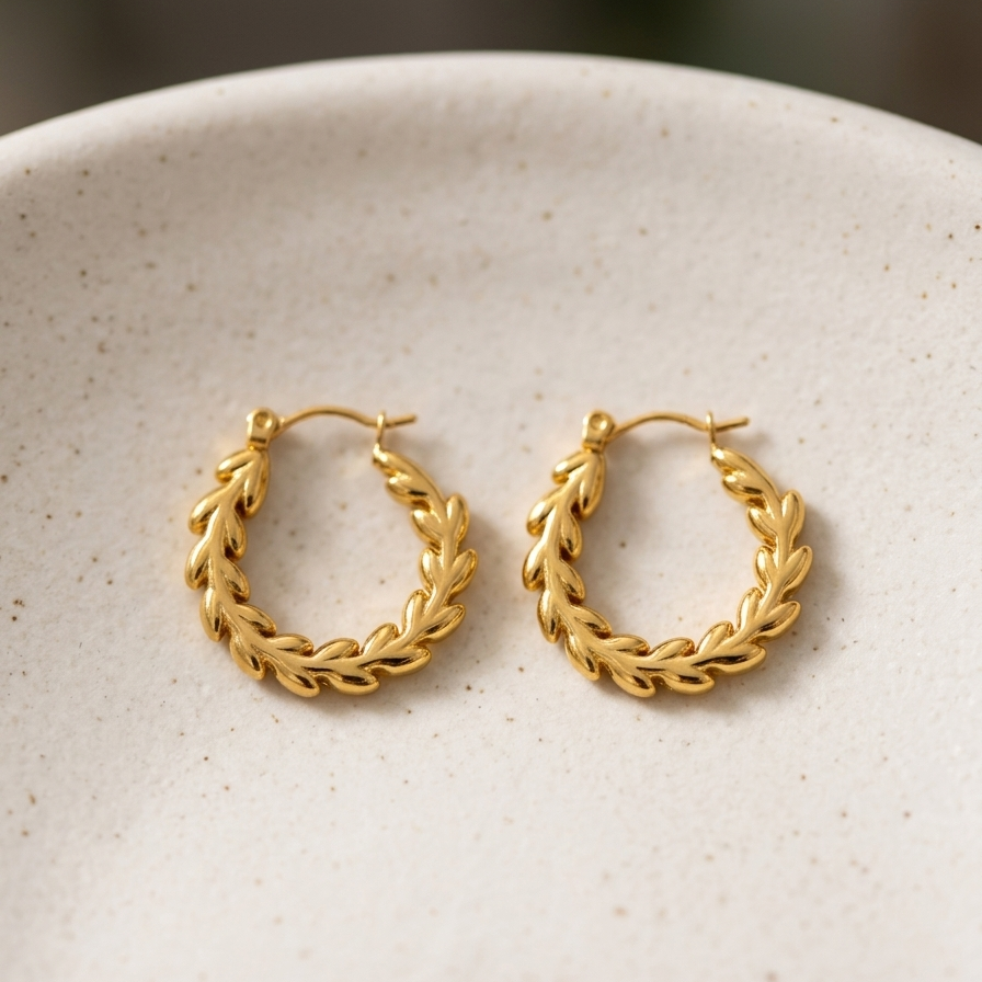 Close up of 18k gold plated hoop earrings with an intricate braided vine and leaf texture, sitting on a matte white plate.