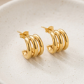 Pair of 18k gold plated hoop earrings featuring a stacked, triple-band design, resting on a beige ceramic plate.