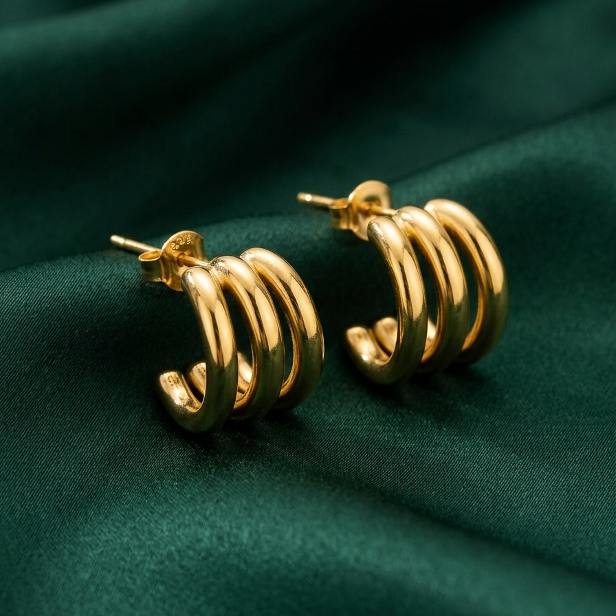 Pair of 18k gold plated hoop earrings featuring a stacked, triple-band design, resting on a green silk fabric.