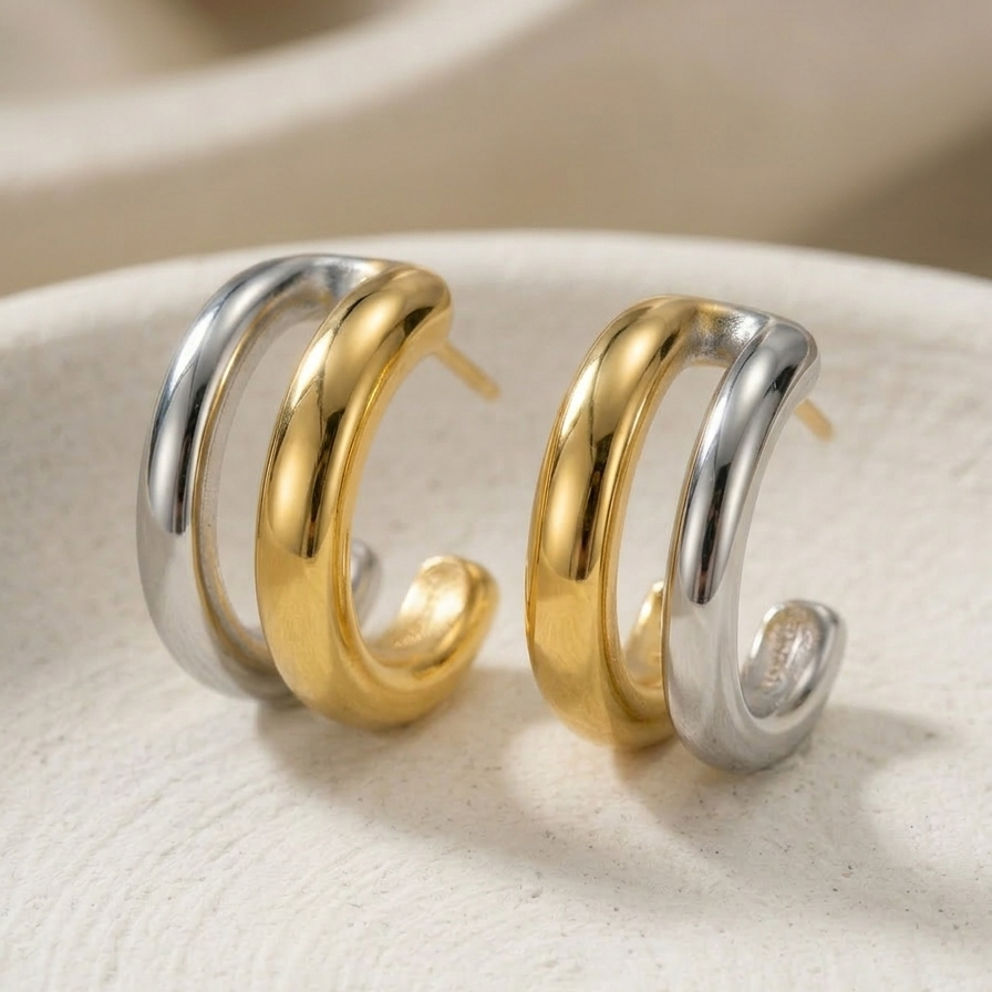 Close up of small double hoop earrings, featuring one gold band and one silver band intertwined, resting on a textured white surface.
