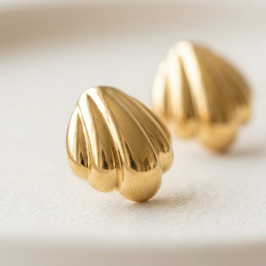 Close up of 18k gold plated stud earrings shaped like polished minimalist scallop seashells, resting on a matte white ceramic dish.