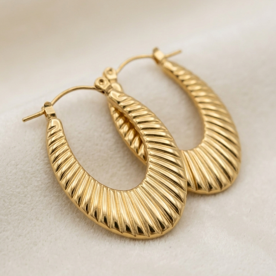 Close of a 18k gold plated elongated oval hoop earrings featuring a ribbed croissant texture, resting on a matte white plate.