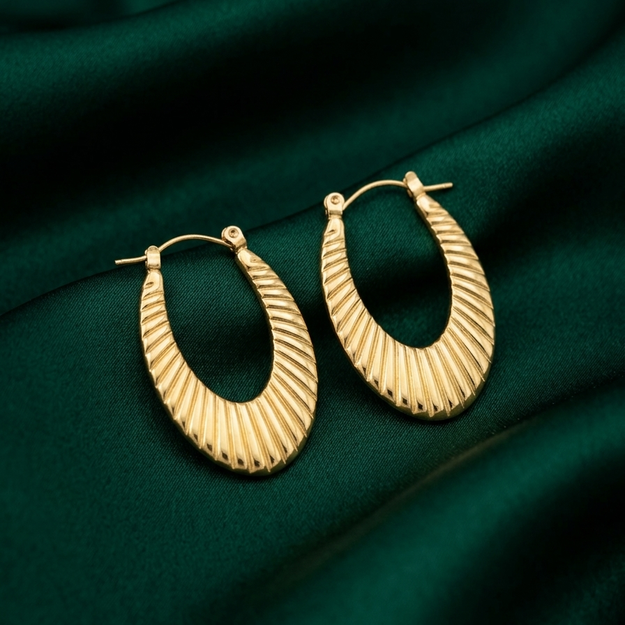 18k gold plated elongated oval hoop earrings featuring a ribbed croissant texture, resting on a matte white plate.on a green fabric background