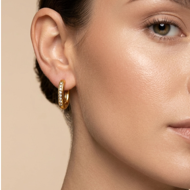 Close-up of a woman wearing Pair of 18k gold plated oval huggie hoop earrings featuring a center row of sparkling CZ diamonds. against a beige background