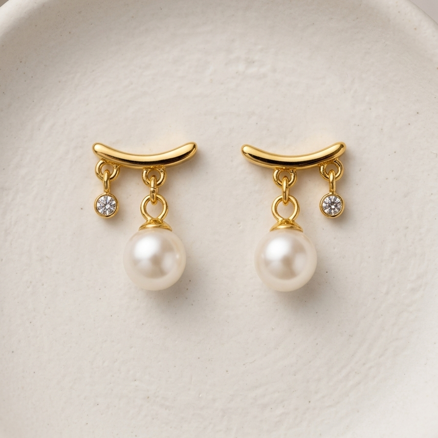 18k gold plated stud earrings featuring a curved gold bar with a dangling white pearl and a smaller dangling CZ diamond, on a white ceramic surface.