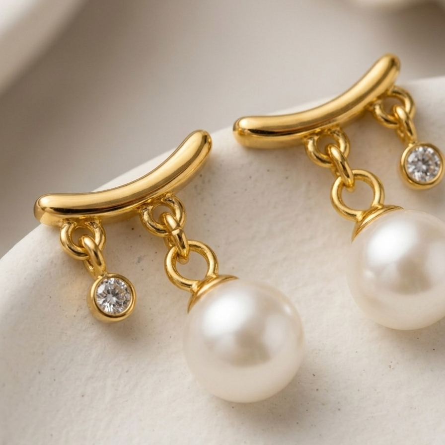 Close up of a 18k gold plated stud earrings featuring a curved gold bar with a dangling white pearl and a smaller dangling CZ diamond, on a white ceramic surface.