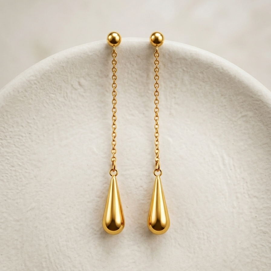 A pair of gold dangle earrings with a ball stud, short chain, and teardrop drop, displayed on a textured white ceramic plate.