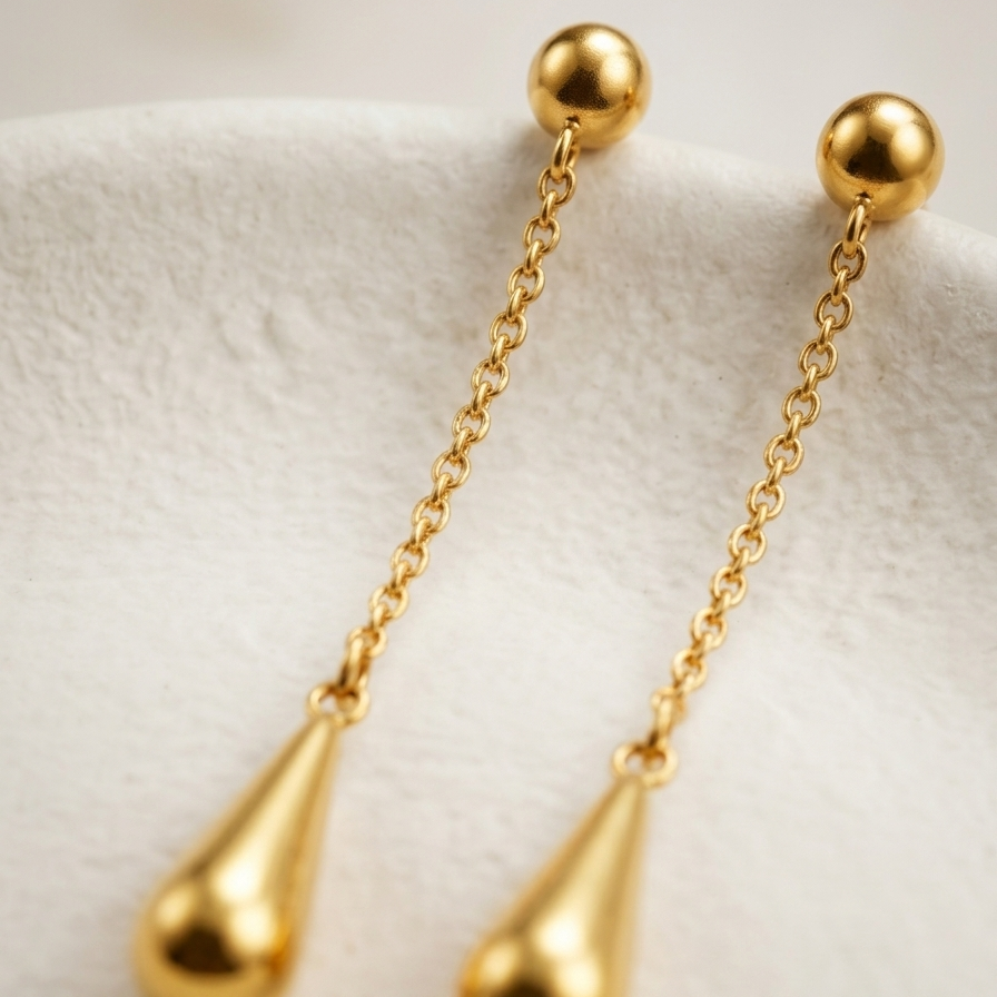 A pair of gold dangle earrings with spherical studs, delicate chain links, and teardrop-shaped drops, displayed on a soft white textured surface.
