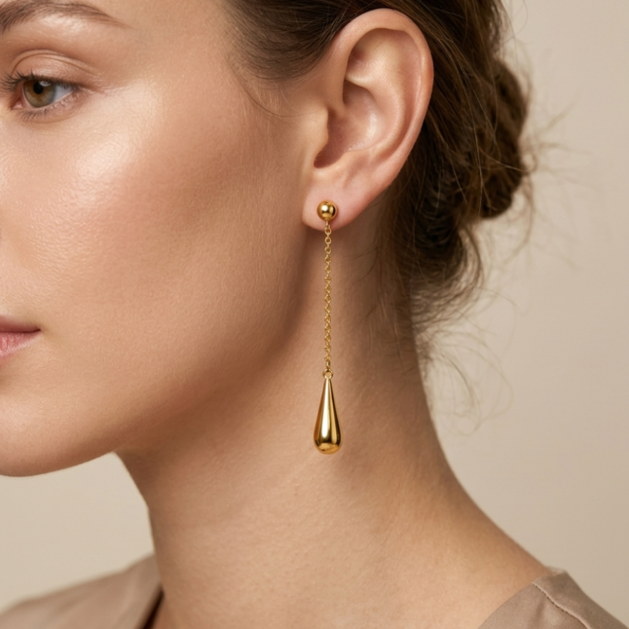 A close-up of a woman's ear and neck wearing a delicate gold dangle earring with a small stud and a teardrop-shaped drop, against a soft beige background.