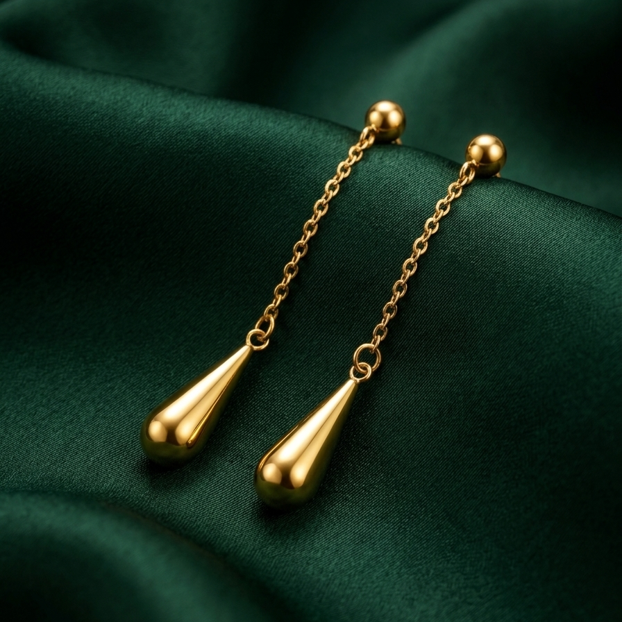 A pair of gold dangle earrings with teardrop-shaped drops and small bead details, displayed on a dark green satin fabric background.