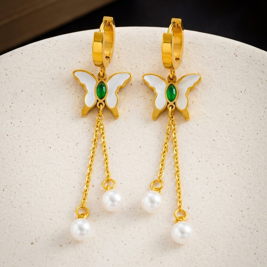 Butterfly earrings with gold frames, green gemstones, and pearl drops on a beige surface.