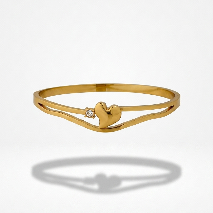 A gold double-band bangle bracelet featuring a polished heart charm in the center and a small clear gemstone on the left band, shown against a white background with a soft shadow below.