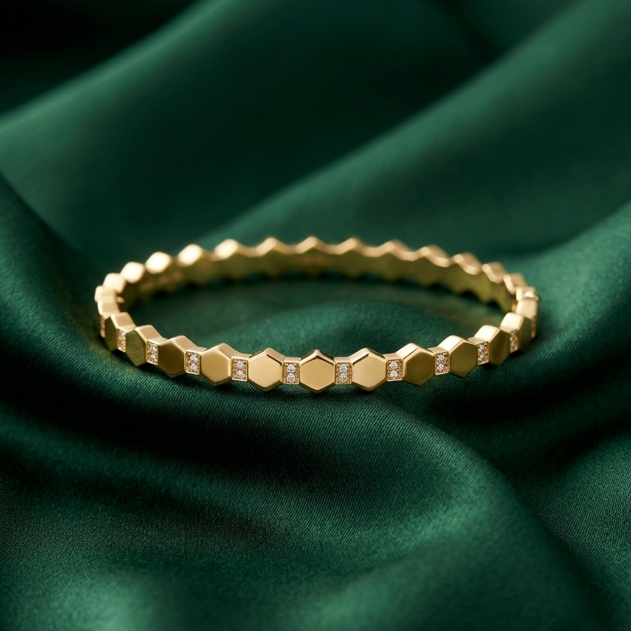 A gold hexagonal beaded bracelet with small clear stones, resting on draped dark green satin fabric.