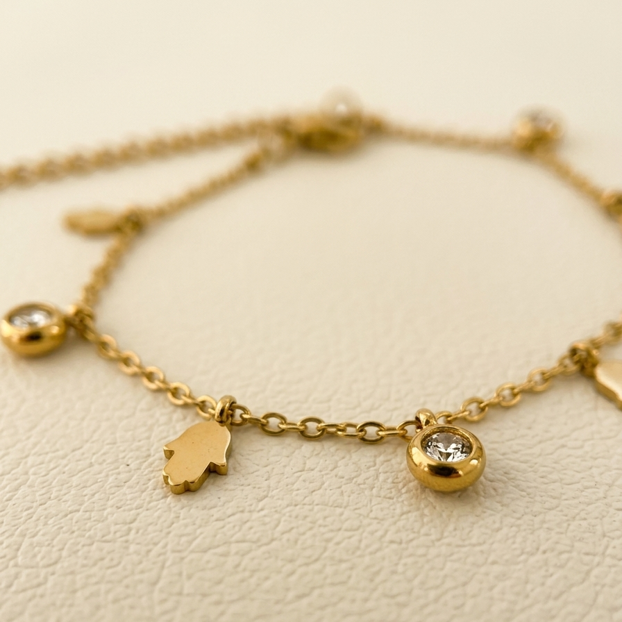 A delicate gold chain bracelet featuring a small hamsa hand charm and a round bezel-set clear crystal charm, displayed on a textured cream surface.