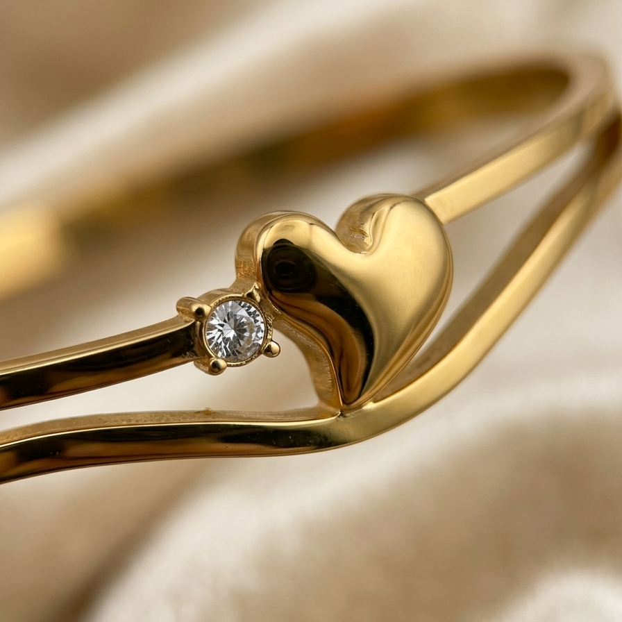 A close-up of a gold bangle bracelet featuring a polished heart-shaped centerpiece and a small round clear gemstone, set against a soft beige fabric background.