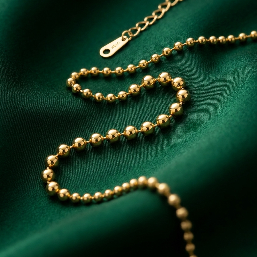 A gold ball chain necklace with a lobster clasp, elegantly draped on a textured dark green fabric background.