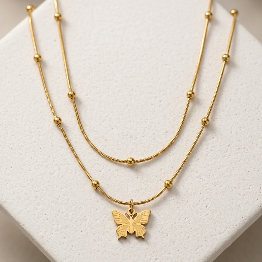 A delicate double-layered gold necklace with small bead accents and a butterfly pendant, displayed on a textured white jewelry stand.