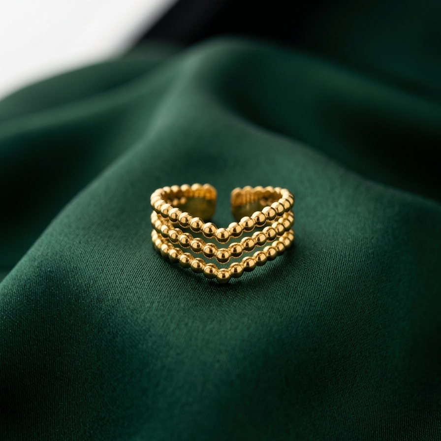 A gold-toned three-band beaded ring resting on draped dark green fabric.