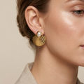 A close-up of a woman's ear wearing a gold fan-shaped stud earring with a white pearl at the top, against a soft beige background.