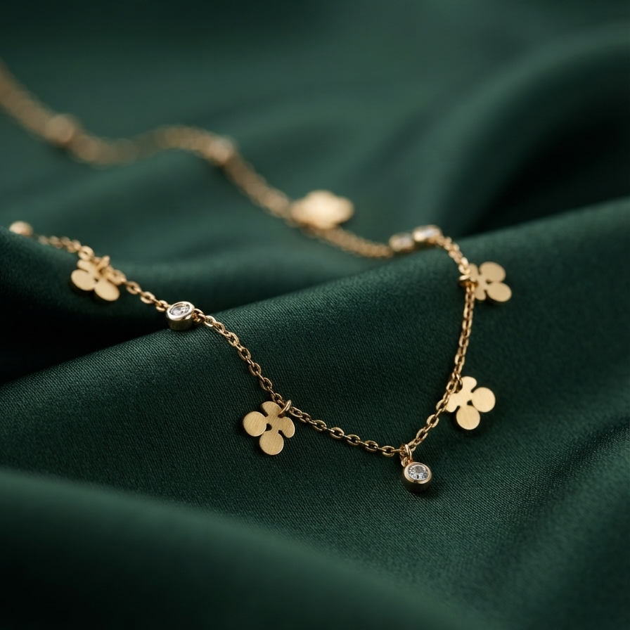 A delicate gold chain necklace with small flower charms and a single clear stone pendant, displayed on draped dark green satin fabric.