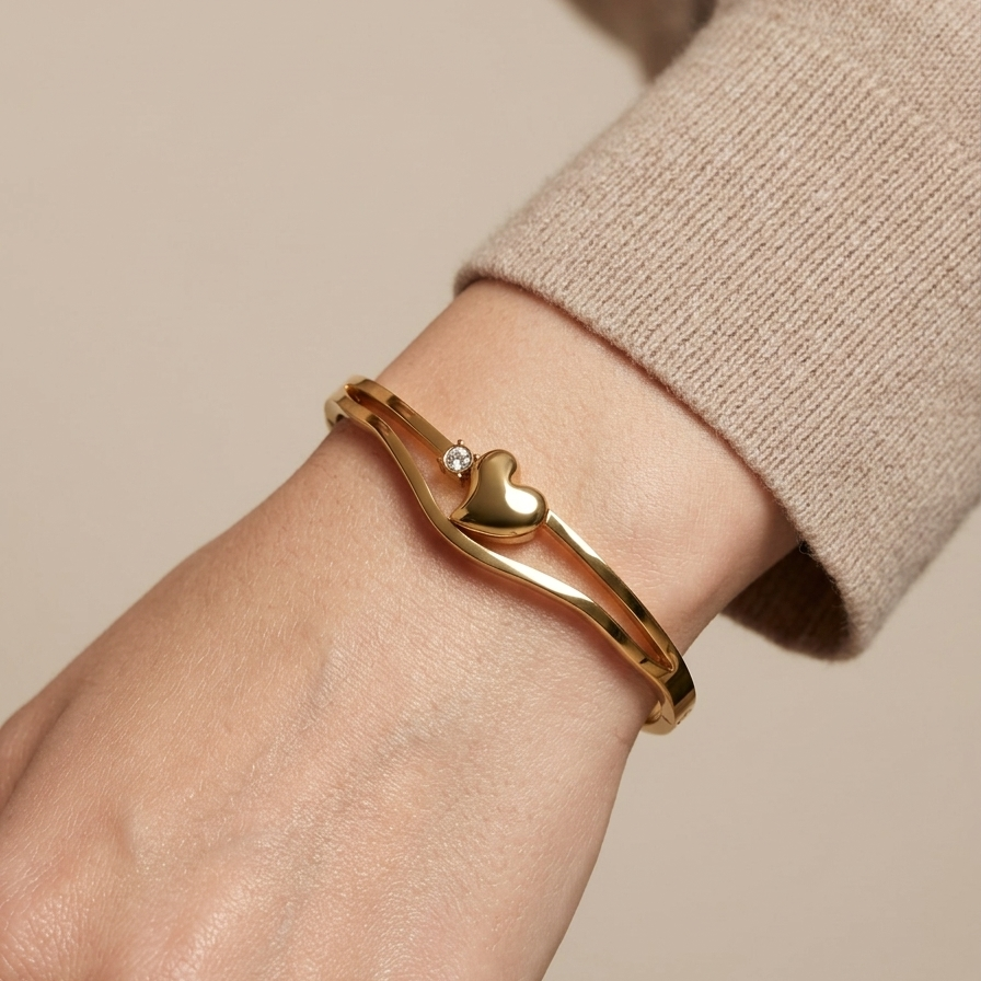 A gold-plated bangle bracelet with a small heart charm and a tiny clear stone, shown worn on a person's wrist with a beige knit sweater cuff visible.