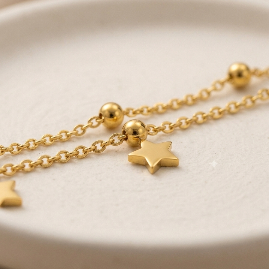 A close-up of a delicate gold chain bracelet with small gold beads and star-shaped charms, resting on a textured off-white ceramic dish.