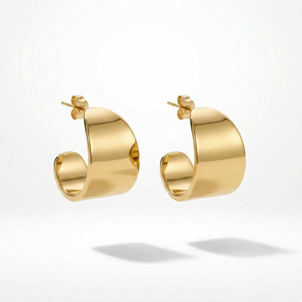 Wide sculpted gold huggie earrings on beige stone background.