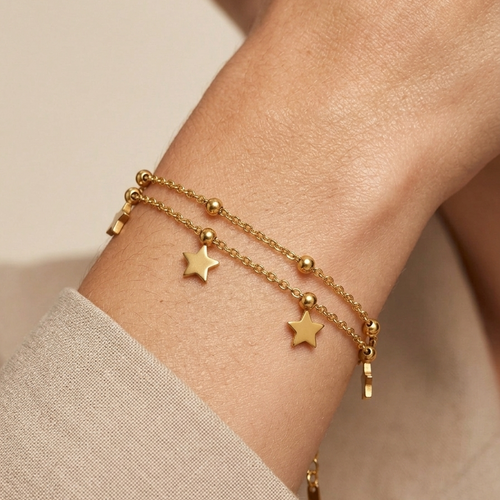 A close-up of a person's wrist wearing two delicate gold chain bracelets, each adorned with small star charms and bead details, paired with a beige sleeve.