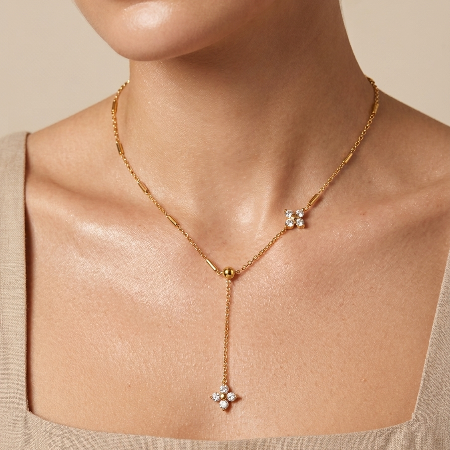 A gold Y-necklace with three floral starburst motifs, worn by a model in a beige linen top against a warm beige background.