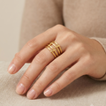 A close-up of a hand wearing a gold ring with three parallel horizontal bands, styled with a beige knit sweater against a neutral beige background.