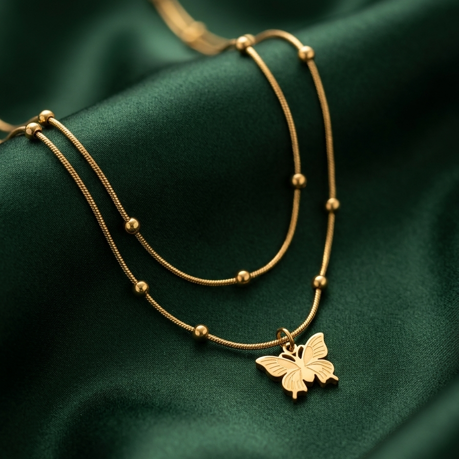 A gold double-layered necklace with small bead accents and a butterfly pendant, displayed on a draped dark green satin fabric background.