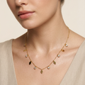 A close-up of a model wearing a delicate gold leaf necklace with small clear crystal accents, paired with a beige linen top against a warm beige background.