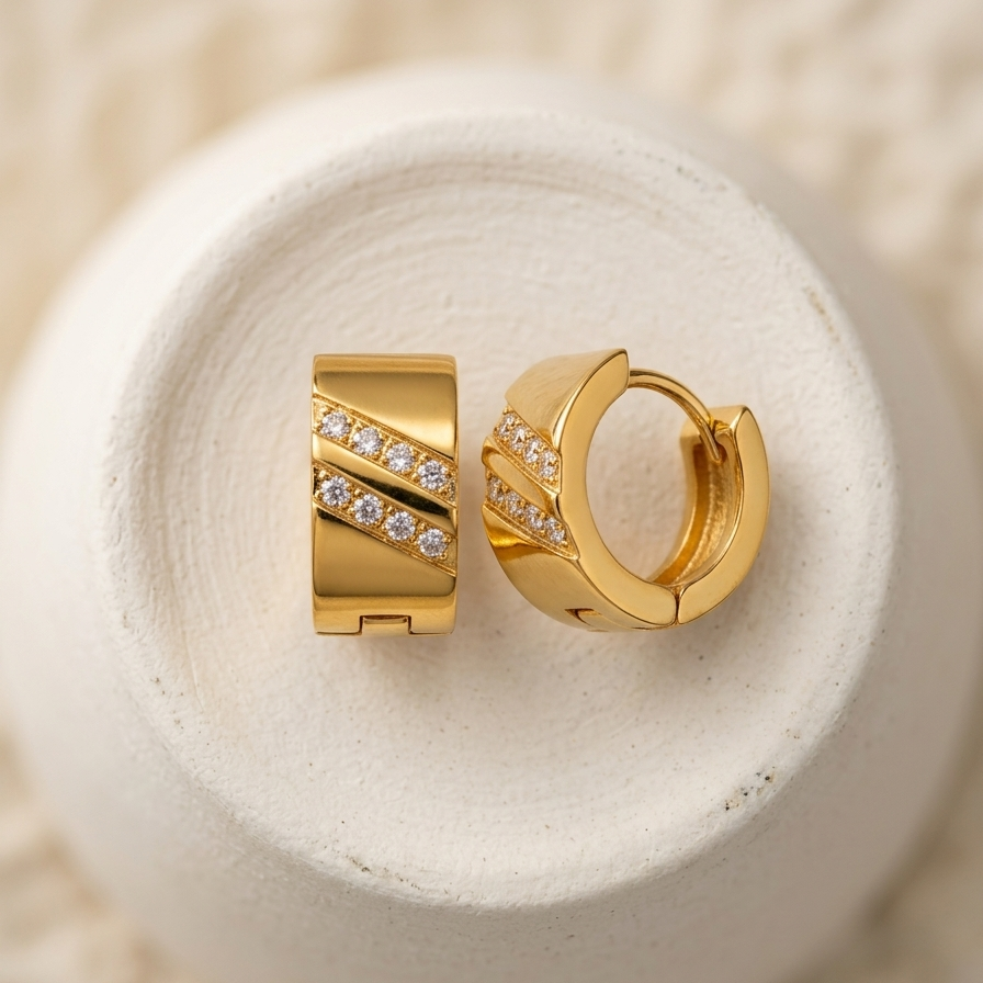 A pair of gold huggie hoop earrings with diagonal rows of clear pavé stones, displayed on a textured white ceramic dish over a soft beige knit fabric background.