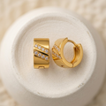 A pair of gold huggie hoop earrings with diagonal rows of clear pavé stones, displayed on a textured white ceramic dish over a soft beige knit fabric background.