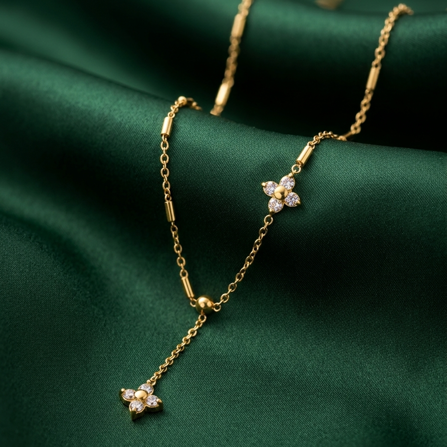 A delicate gold Y-necklace with two small flower-shaped clusters of clear stones, draped elegantly on a dark green satin fabric background.