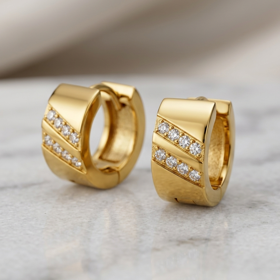 A pair of gold huggie hoop earrings with diagonal rows of pavé-set clear stones, displayed on a light marble surface with a soft beige fabric background.
