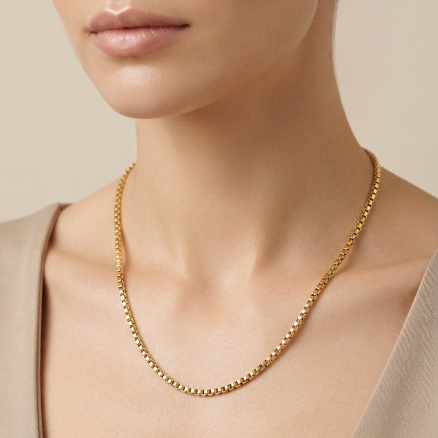 A close-up of a model wearing a delicate gold beaded chain necklace, styled with a beige top against a soft beige background.