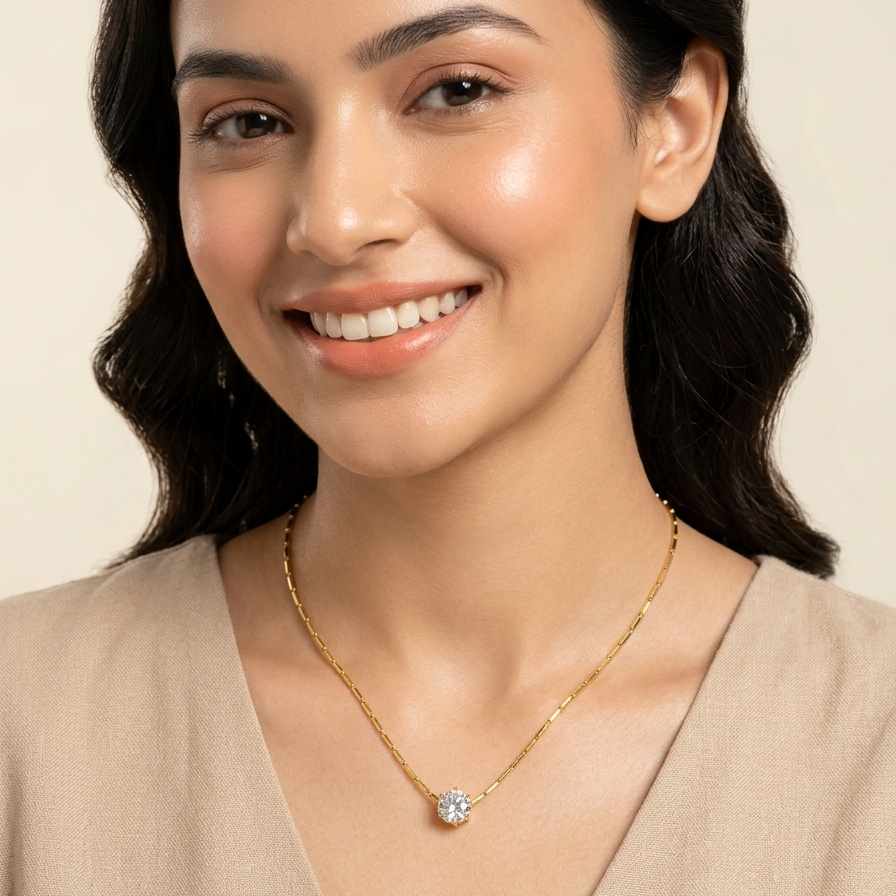 A smiling woman with dark hair models a delicate gold chain necklace with a small, clear, round-cut gemstone pendant. She is wearing a beige V-neck top against a solid, warm off-white background.