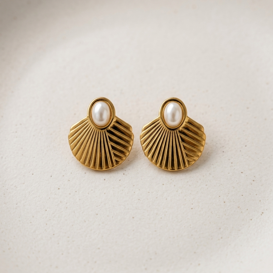 A pair of gold shell-shaped stud earrings with a white pearl at the center, displayed on a light beige textured ceramic plate.