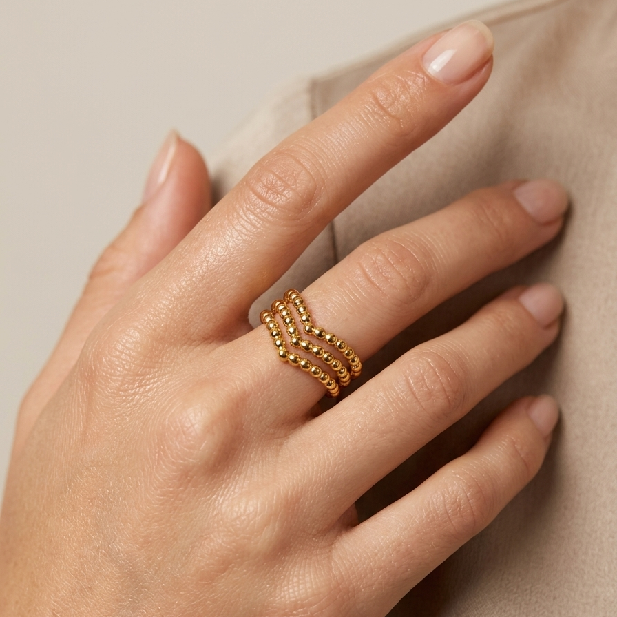 A close-up of a hand wearing a delicate gold ring with three parallel rows of small round beads, resting against a soft beige fabric background.