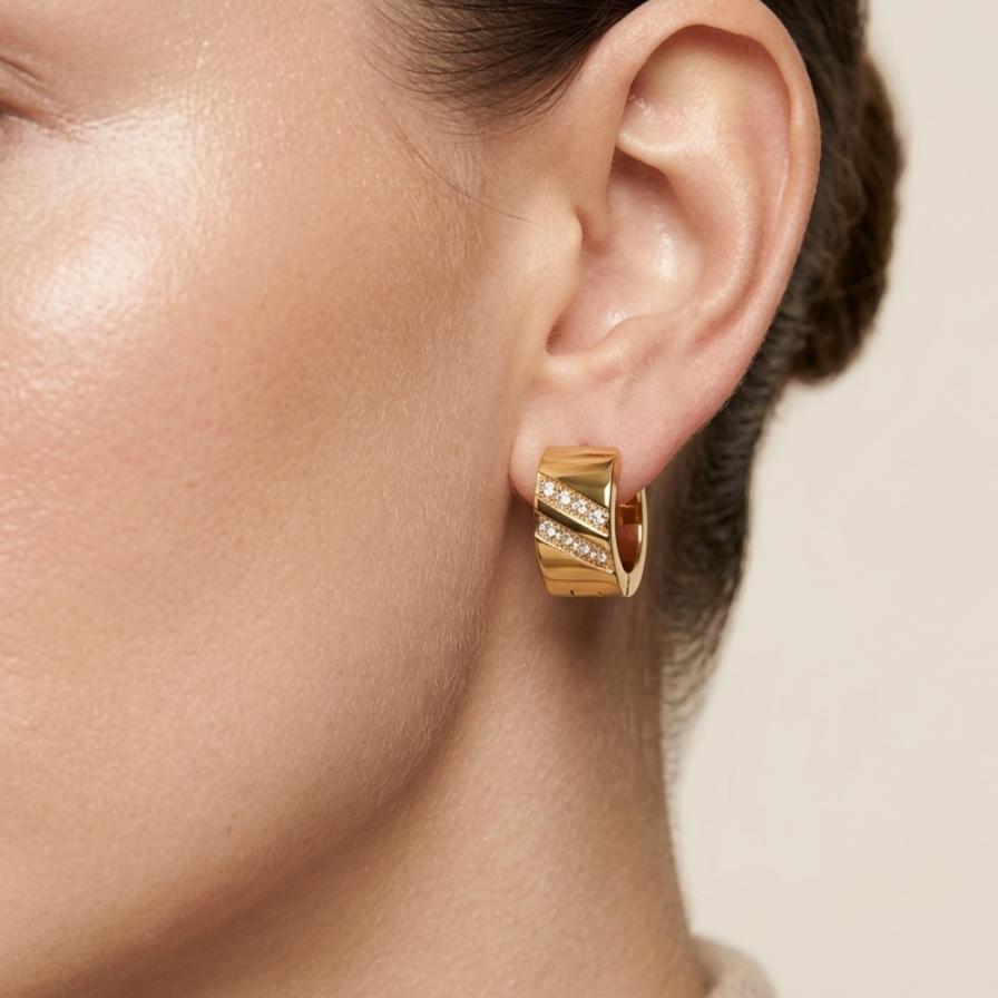 A close-up of a model's ear wearing a gold huggie hoop earring with diagonal channel-set clear stones, set against a soft beige background.