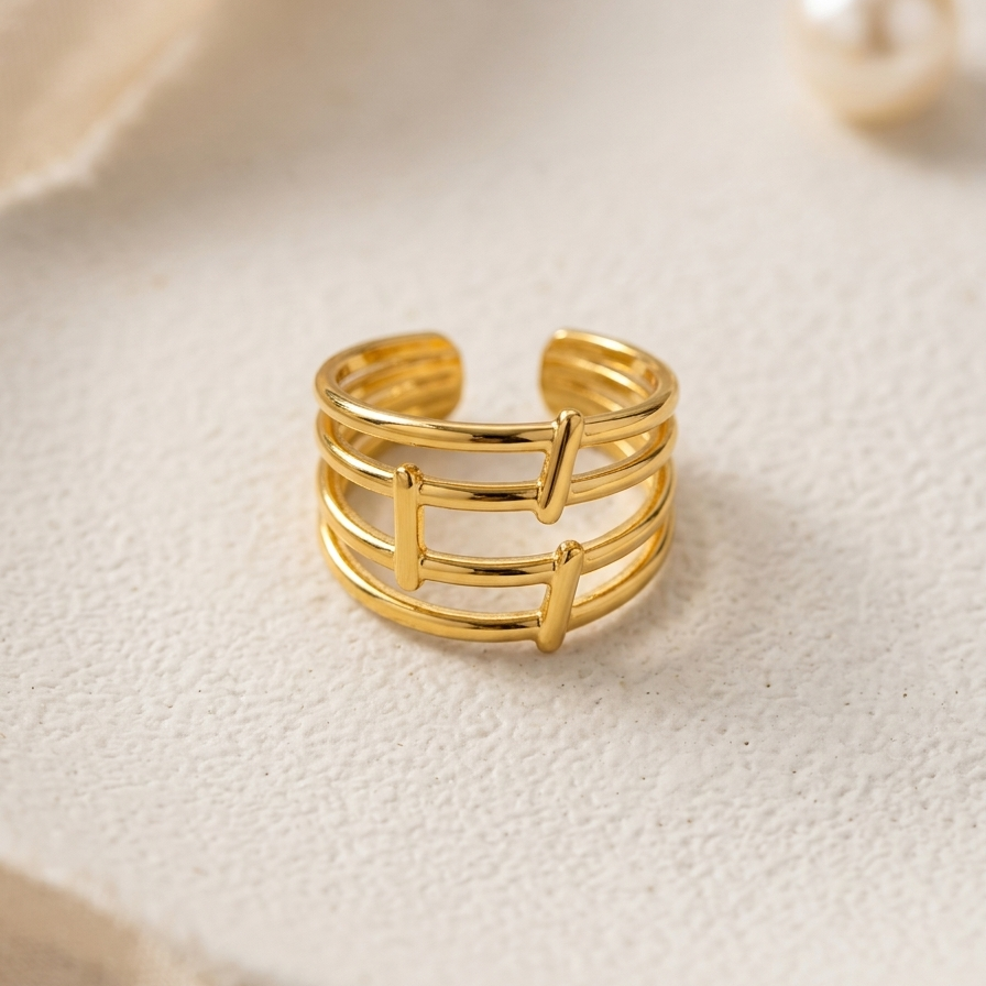 A gold-plated open bangle ring with a geometric, multi-strand bar design, displayed on a textured cream-colored surface next to a pearl.