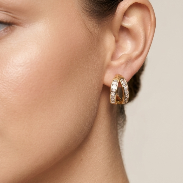 A close-up of a woman's ear wearing a small gold hoop earring adorned with clear stones, set against a soft beige background.