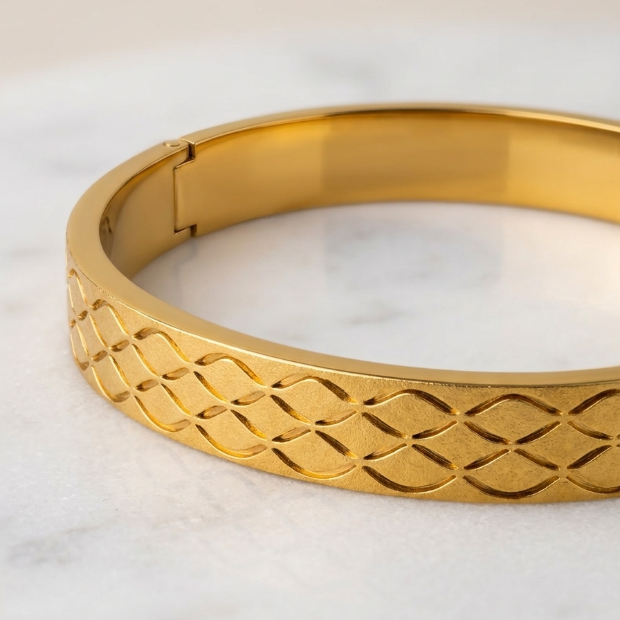A gold-toned metal bangle bracelet with an open clasp and a textured, woven geometric pattern on the outer band, resting on a light marble surface.