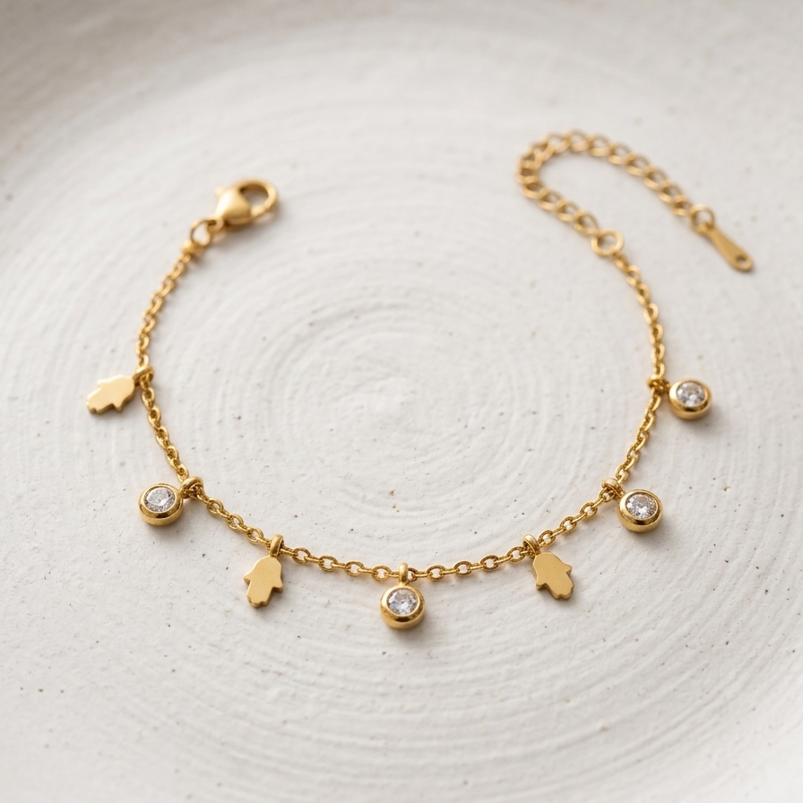 A delicate gold chain bracelet with five small hanging charms, including three hand-shaped charms and two round bezel-set clear stones, displayed on a textured white ceramic plate.