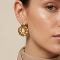 A close-up of a woman wearing a textured gold hoop earring with a twisted, spiral design. She is wearing a beige high-neck top and a matching beige scarf against a solid beige background.