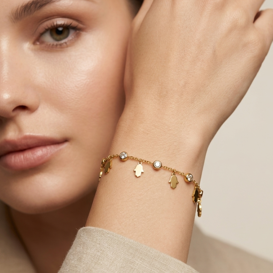 A close-up lifestyle shot of a model wearing a delicate gold charm bracelet on her wrist. The bracelet features small gold charms, including a Star of David and a small heart, with a few clear stones.