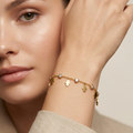 A close-up lifestyle shot of a model wearing a delicate gold charm bracelet on her wrist. The bracelet features small gold charms, including a Star of David and a small heart, with a few clear stones.
