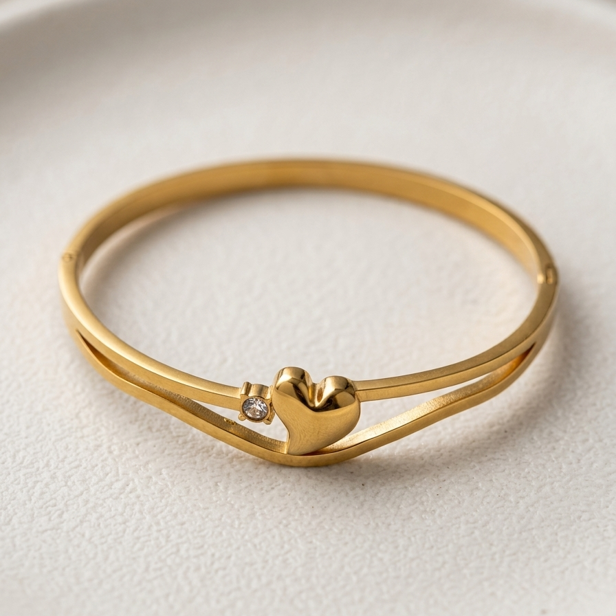 A gold-plated double-band ring featuring a small polished heart charm in the center with a tiny clear accent stone, resting on a textured white ceramic plate.